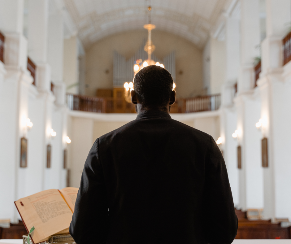 Black man with back towards camera facing an empty congregation in a church