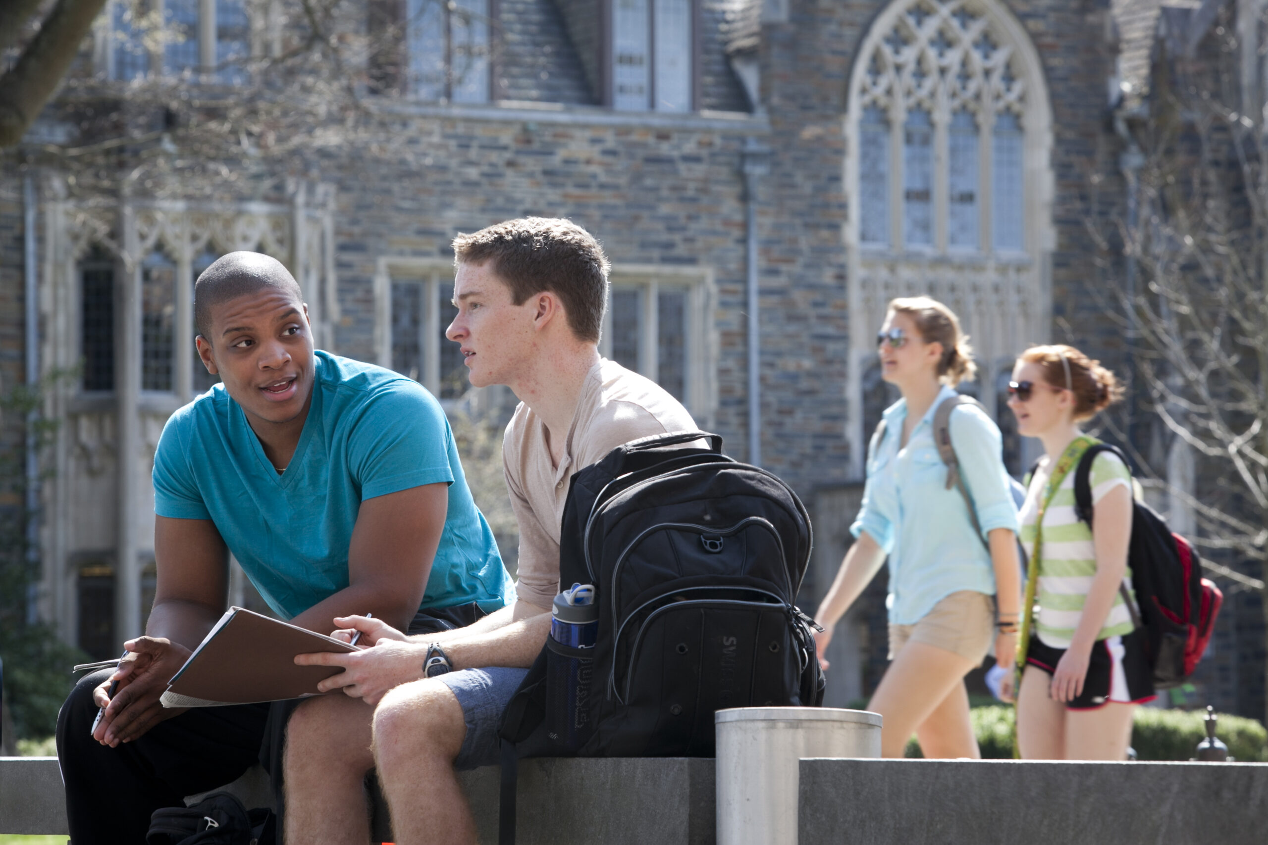 Two young men sitting on a bench and having a conversation with students walking in the background and Duke's campus in the background